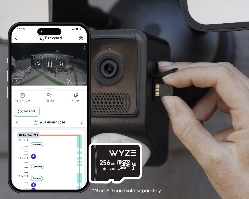 Person inserting a microSD card into a Wyze security cam. The phone in front is displaying a live feed from the cam, which shows a backyard at night.