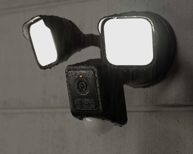 A black security camera with two floodlights attached, mounted to a wall. Rain is falling on the camera and lights.