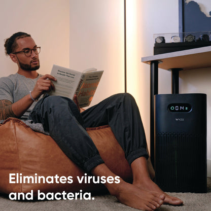 Man reading a book nearby a watch collection and Wyze Air Purifier. White text overlay that says eliminates viruses and bacteria.