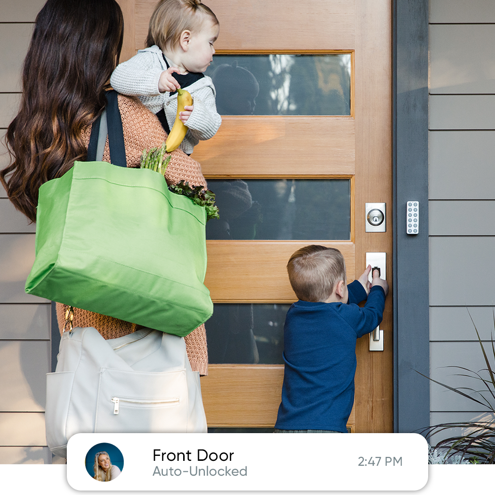 A mother carrying a child while watching her other child open the door to the house.