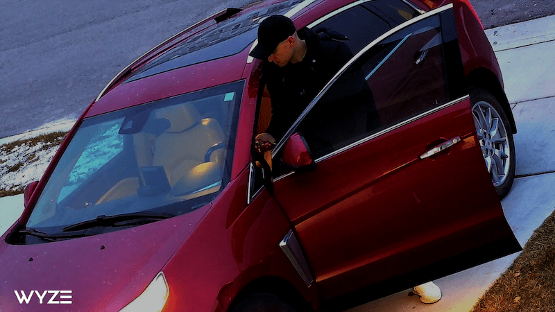 Color night vision of a red car in a driveway with a male driver wearing a black cap, black hoodie, and white shoes.