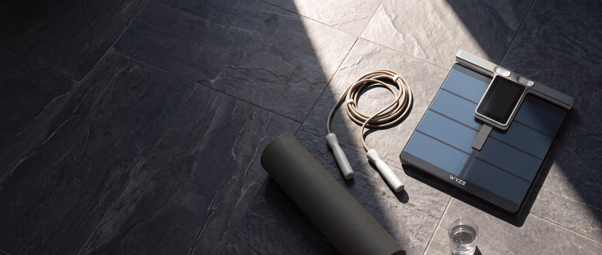 A fitness floor scale with a smartphone resting on top, shown alongside a rolled exercise mat and a jump rope on dark gray stone-look floor tiles.