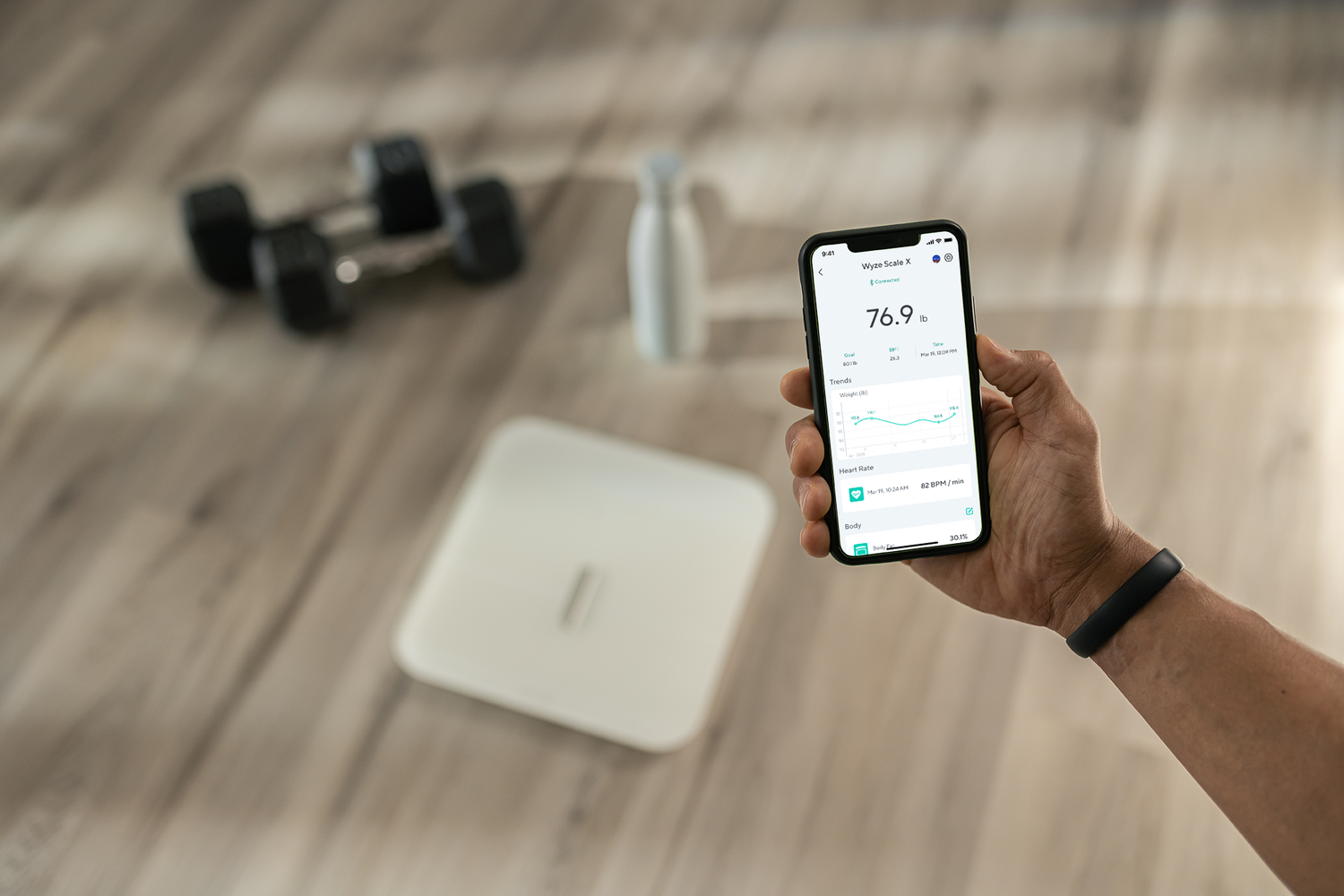 Person holding phone with Wyze app open to scale data. White Scale x, weights, and water bottle in background