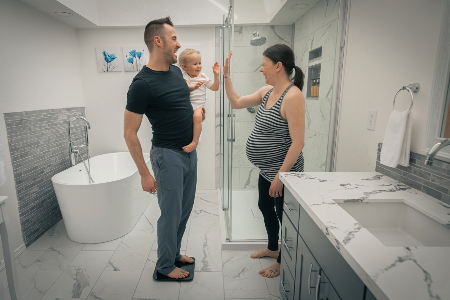 A man and a woman standing in a bathroom, the man is holding a child and they are all on a scale.