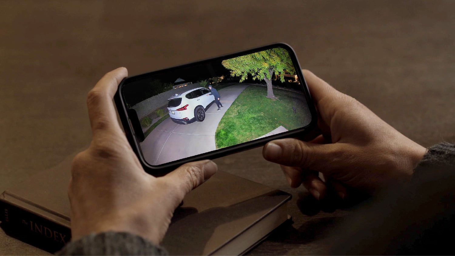 Hands holding phone with livestream of man approaching car parked in a driveway. Security camera light illuminates the scene.