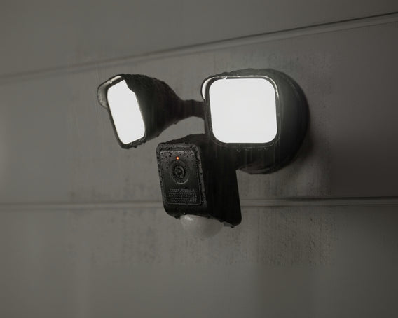 A black security camera with two floodlights attached, mounted to a wall. Rain is falling on the camera and lights.