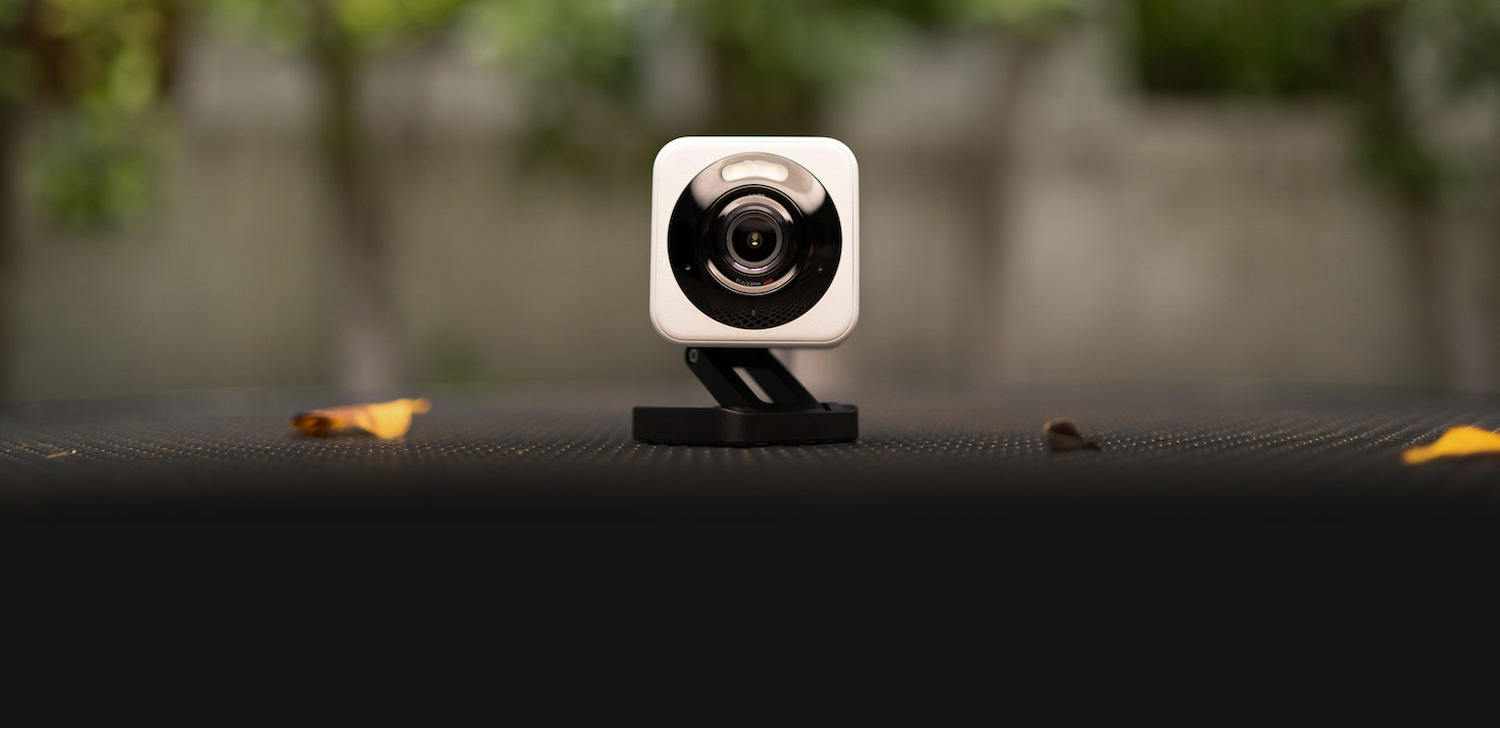 A small square security camera with a white body and black lens surround mounted on a black stand, photographed outdoors against a blurred green and gray background.