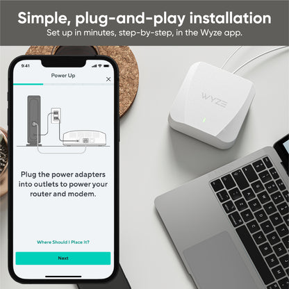 Wyze Mesh Router on a desk beside a laptop with a superimposed phone screen showing part of the setup process in the Wyze app.